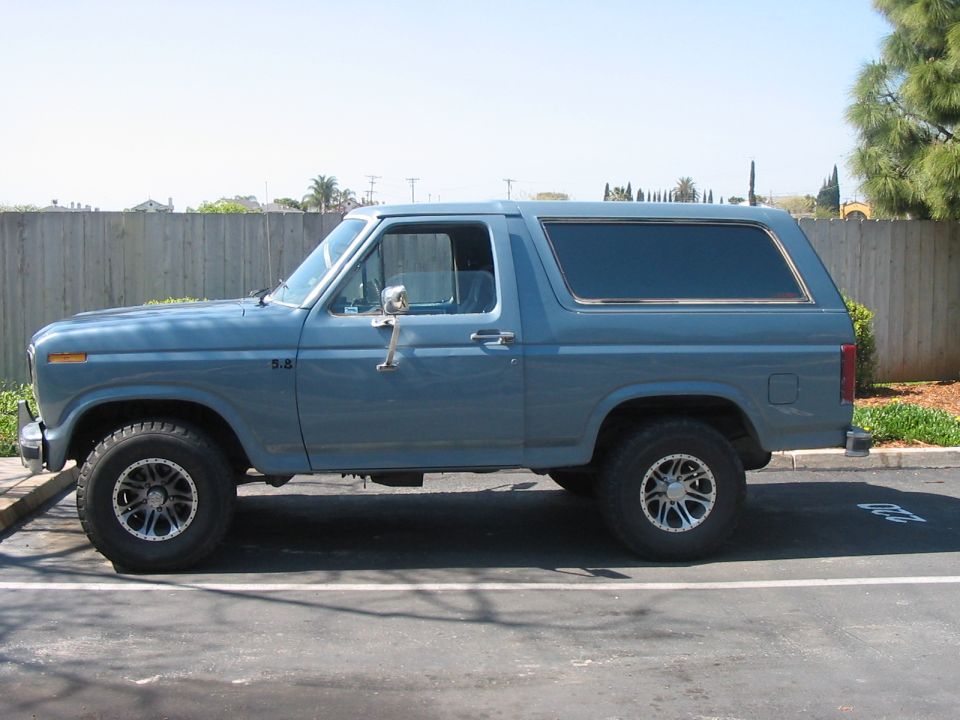 Light blue 1986 Ford Bronco side view with aftermarket alloy wheels, parked by a wooden fence
