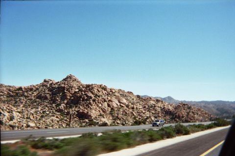 1990 Ford Bronco - lone pickup truck travels a divided desert highway below a rugged boulder-strewn hillside under clear blue sky.