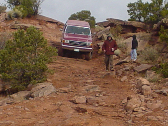 Burgundy full-size Ford van descending a rocky desert 4x4 trail with two onlookers nearby