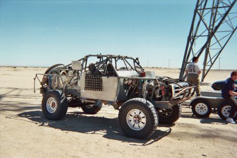 1990 Ford Bronco - skeletal tube-chassis off-road race car sits on the desert next to a trailer as two people stand nearby.