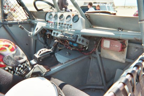 1990 Ford Bronco - stripped interior of a race buggy reveals a metal dash with gauges, toggle switches and exposed wiring beside helmets.
