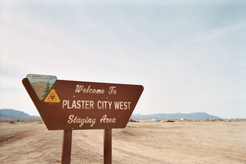 1990 Ford Bronco - brown wooden sign reads 'Welcome To Plaster City West Staging Area' on an open desert flat under hazy sky.