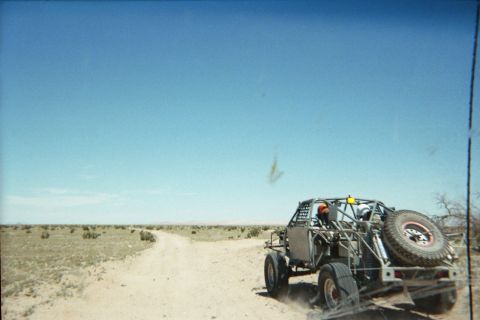 1990 Ford Bronco - tube-frame off-road buggy rides on a trailer kicking up dust along a dirt road across open desert.