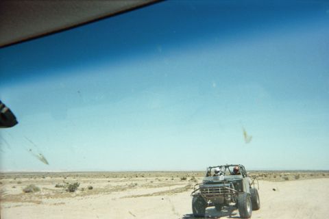 1990 Ford Bronco - Through a speckled windshield, a tube-frame buggy is towed ahead across a flat, sparse desert plain.