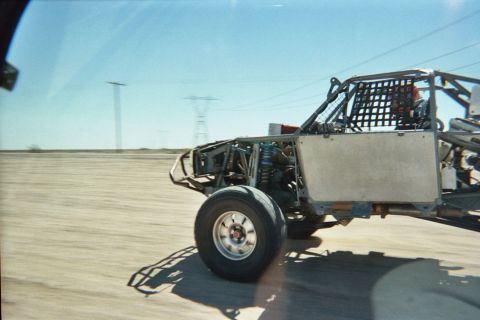 1990 Ford Bronco - tube-frame off-road buggy speeds across desert hardpan with transmission towers and blurred ground behind it.