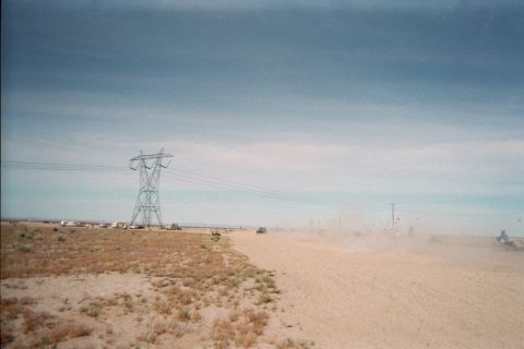 1990 Ford Bronco - steel transmission tower stands over a desert staging area as vehicles raise dust along a sandy track.