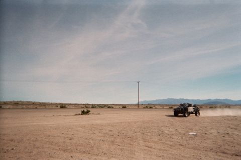 1990 Ford Bronco - small off-road buggy trails dust across a vast flat desert next to a lone power pole under a wispy sky.