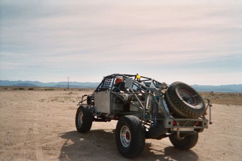 1990 Ford Bronco - tube-chassis off-road buggy carrying a rear spare tire sits on open desert ground beneath a soft cloudy sky.