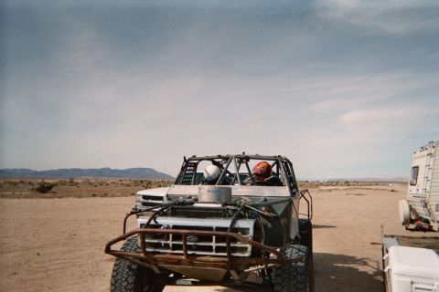 1990 Ford Bronco - white-bodied tube-frame buggy with two helmeted occupants idles on the desert next to a parked RV.