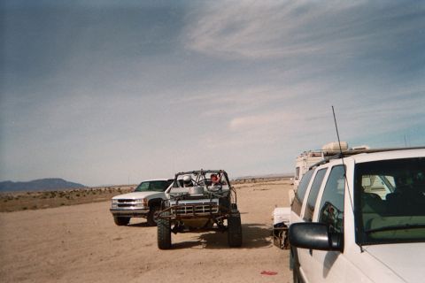 1990 Ford Bronco - white pickup truck, a tube-frame off-road buggy and a white van sit parked together on an open desert flat.