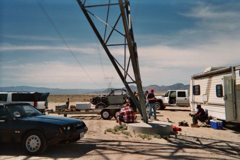1990 Ford Bronco - People gather at a desert camp beneath a transmission tower, with a Mustang, trailers, an RV and an off-road buggy nearby.