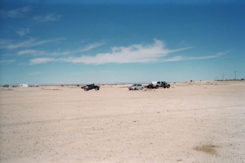 1990 Ford Bronco - pickup, a car and another truck are parked in the distance across a broad pale desert flat under a clear sky.