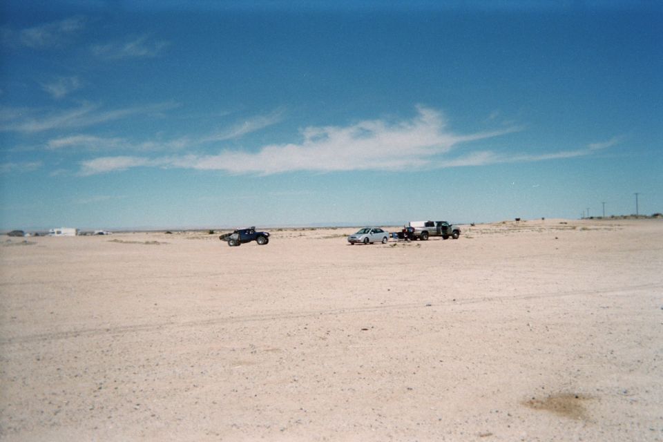 Two pickups and a car parked far across a wide pale desert flat under blue sky