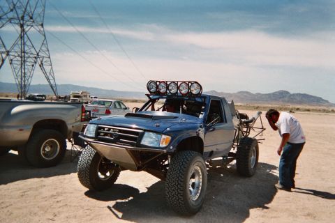 1990 Ford Bronco - blue off-road prerunner pickup with a roof light bar and long-travel suspension sits in the desert as a man inspects it.