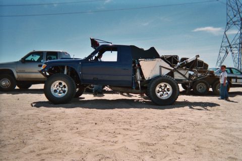 1990 Ford Bronco - blue open-cockpit off-road race buggy on oversized tires sits on desert sand beside a silver pickup truck under a clear sky.