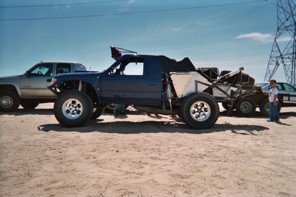 Blue tube-frame off-road buggy on large tires parked on desert sand near a pickup