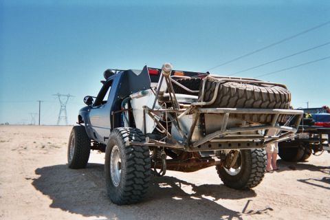1990 Ford Bronco - Front view of a tube-frame desert off-road buggy on large knobby tires with a spare tire mounted at the rear, parked on sand.