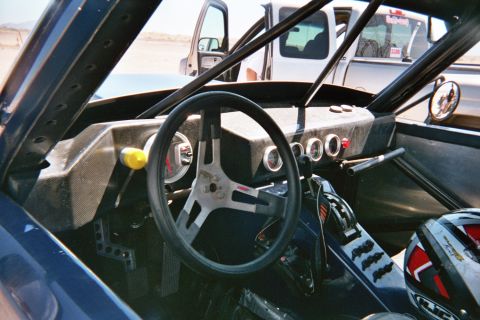1990 Ford Bronco - Interior of an off-road race vehicle with a black steering wheel, round gauges, carbon-look dash and a roll cage; a helmet sits at right.