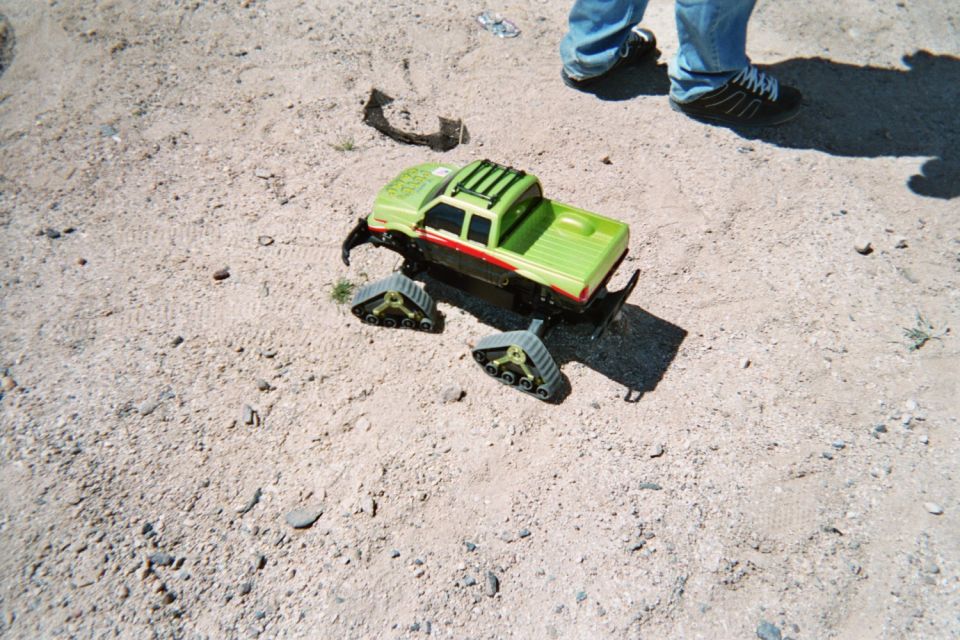 Green toy RC monster truck with tracked wheels sitting on dirt beside a person's feet