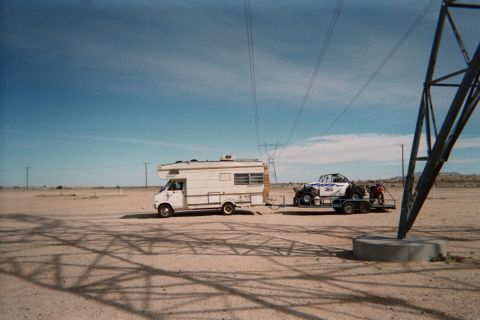 1990 Ford Bronco - white motorhome towing a trailer that carries an off-road buggy parked on desert flats beneath high-tension power lines.