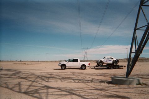 1990 Ford Bronco - white crew-cab pickup tows a flatbed trailer carrying a white off-road buggy across desert flats under power lines and blue sky.