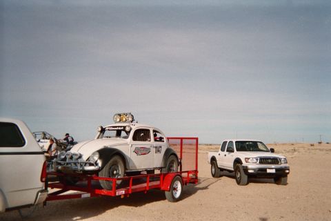 1990 Ford Bronco - white Volkswagen Beetle-based Baja Bug race car with a roof light bar sits on a red trailer next to a white crew-cab pickup in the desert.