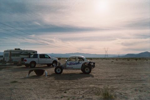 1990 Ford Bronco - white Beetle-based off-road Baja Bug is parked on the open desert at dusk near a white pickup and a motorhome, with mountains behind.