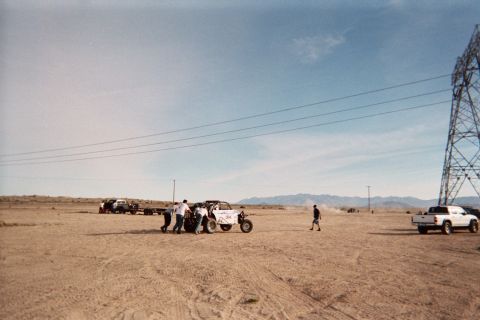 1990 Ford Bronco - group of people push a white off-road buggy across a wide desert flat, with a pickup, trailer and distant mountains in the background.
