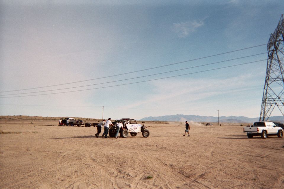 People pushing a white off-road buggy across a wide desert flat with mountains behind