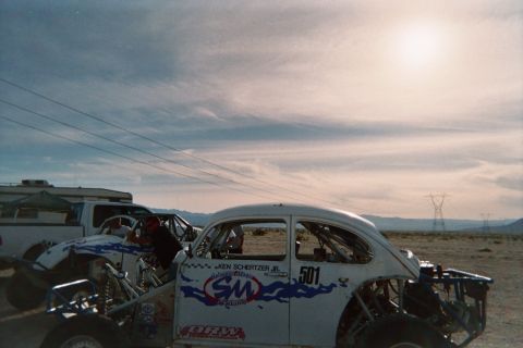 1990 Ford Bronco - white Baja Bug race car numbered 501 with blue flame graphics and 'Ken Schertzer Jr.' lettering sits in the desert at sunset.