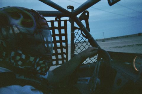1990 Ford Bronco - driver wearing a flame-and-checkered patterned helmet grips the steering wheel inside an off-road buggy at dusk, desert visible ahead.