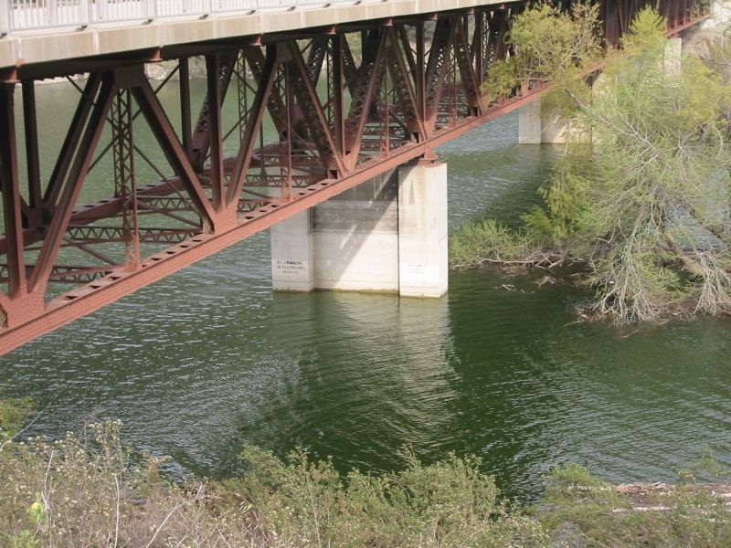 Underside of a red steel truss bridge over green river water with a concrete pier