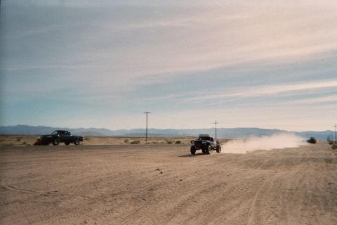 1990 Ford Bronco - off-road buggy kicks up a cloud of dust on a desert road while a pickup truck waits at left, with mountains along the horizon.