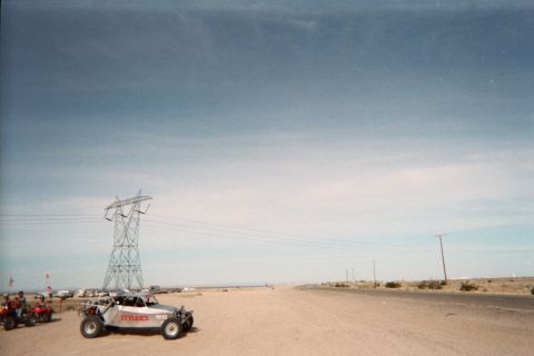 1990 Ford Bronco - white off-road buggy parked beside a high-tension power-line tower on a vast desert flat, with ATVs and a wide blue sky behind.