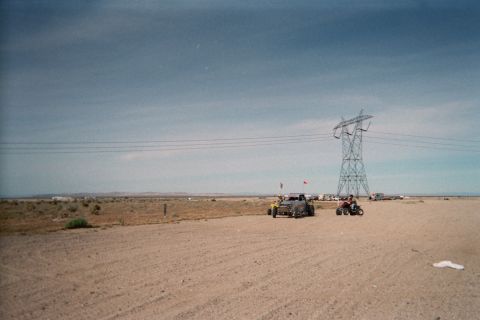 1990 Ford Bronco - Several off-road buggies and ATVs gather near a high-tension power-line tower on open desert flats under a hazy blue sky.