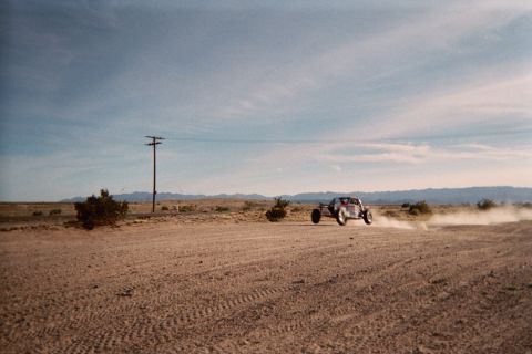 1990 Ford Bronco - off-road buggy races down a sandy desert road throwing up a trail of dust, with a power pole, brush and mountains in the distance.