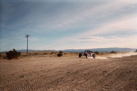 1990 Ford Bronco - off-road buggy speeds along a sandy desert road kicking up dust, passing a wooden power pole with mountains behind under a blue sky.