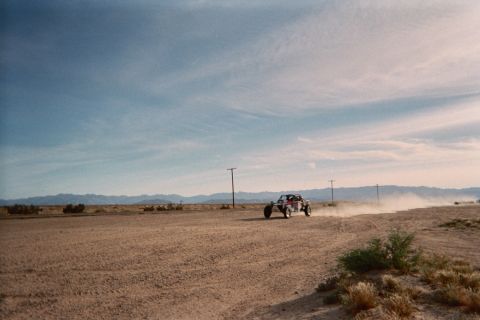 1990 Ford Bronco - open-frame off-road buggy races across a flat desert plain, trailing a plume of dust beside distant power poles and mountains.