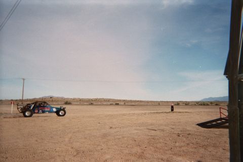 1990 Ford Bronco - blue tube-frame sand rail sits on open desert flats with a wind sock and low mountains in the background.