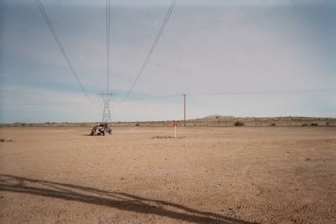 1990 Ford Bronco - small off-road buggy crosses barren desert under sweeping high-voltage transmission lines, with a tire track in the foreground.