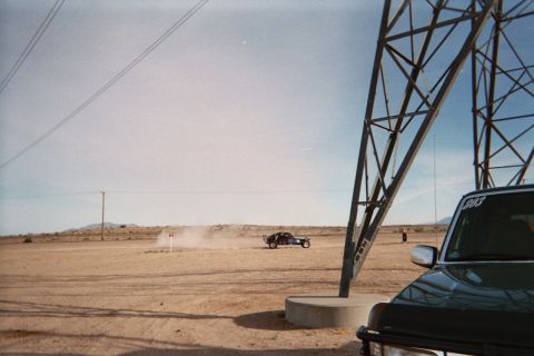 1990 Ford Bronco - steel transmission tower frames a dusty desert flat where an off-road vehicle drives, with a dark pickup parked in the foreground.
