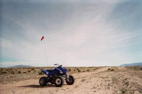 1990 Ford Bronco - blue sport quad ATV with a tall red whip flag sits on a desert two-track with mountains on the horizon.
