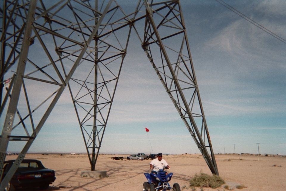 Man on a blue quad ATV beneath a large steel power-line tower in the desert
