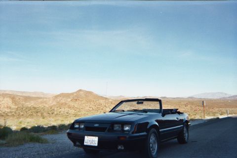 1985 Ford Mustang - A black convertible sits on the side of a road in a desert landscape under a clear blue sky.