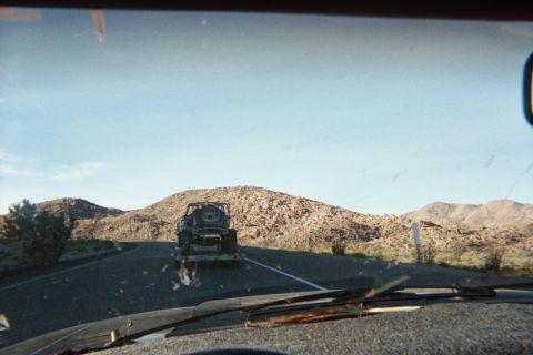 1990 Ford Bronco - Through a wiper-streaked windshield, a boxy Jeep-style vehicle is towed on a trailer along a two-lane desert highway among barren hills.