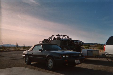 1990 Ford Bronco - dark Ford Mustang convertible with a white top sits in a lot at dusk beside a trailer hauling a tube-frame off-road buggy.