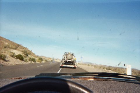1990 Ford Bronco - Through a bug-spattered windshield, a desert highway leads to an off-road vehicle riding on a tow trailer beneath a clear blue sky.