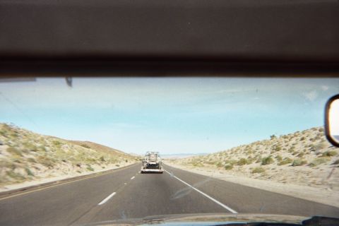 1990 Ford Bronco - Seen through a windshield, a desert highway stretches ahead toward a vehicle being towed on a flatbed trailer between scrubby hills.