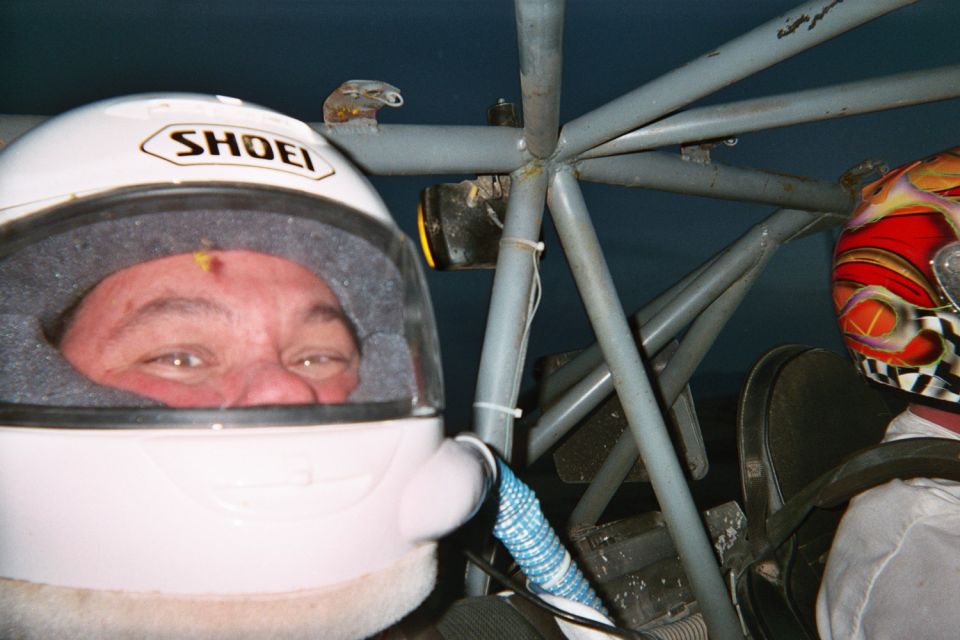 Man in a white Shoei helmet seated in a tube-frame race vehicle beside another driver
