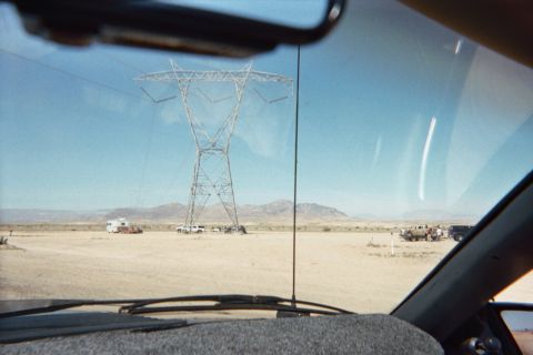 1990 Ford Bronco - Through a windshield, a tall steel transmission tower rises over a desert staging area dotted with parked trucks and a motorhome.